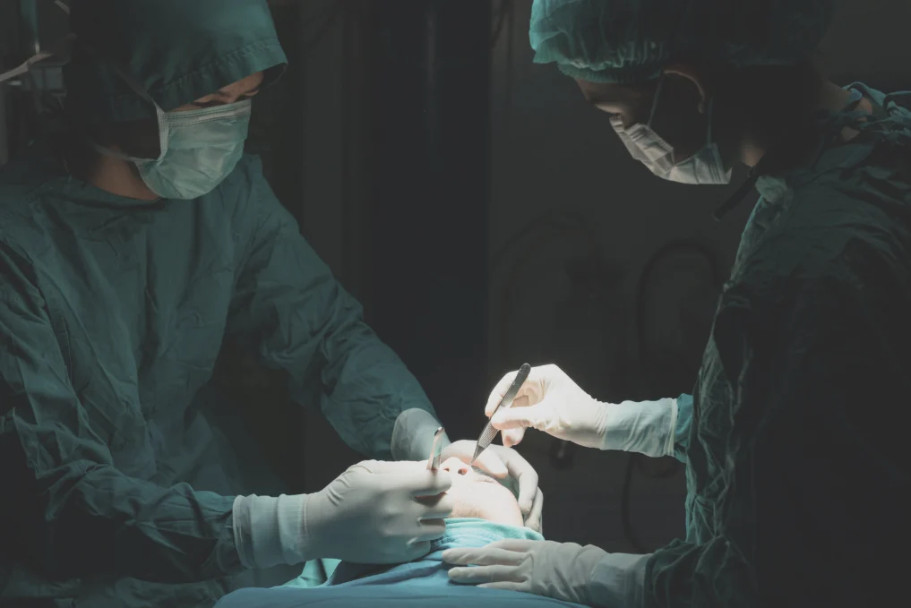 Two surgeons wearing green scrubs, surgical masks, and gloves perform a delicate surgical procedure on a patient's eye area under bright operating room lights.