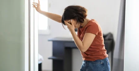 Woman leaning against a doorway holding her head in discomfort, illustrating concerns over whether can dizziness be a sign of eye problems.