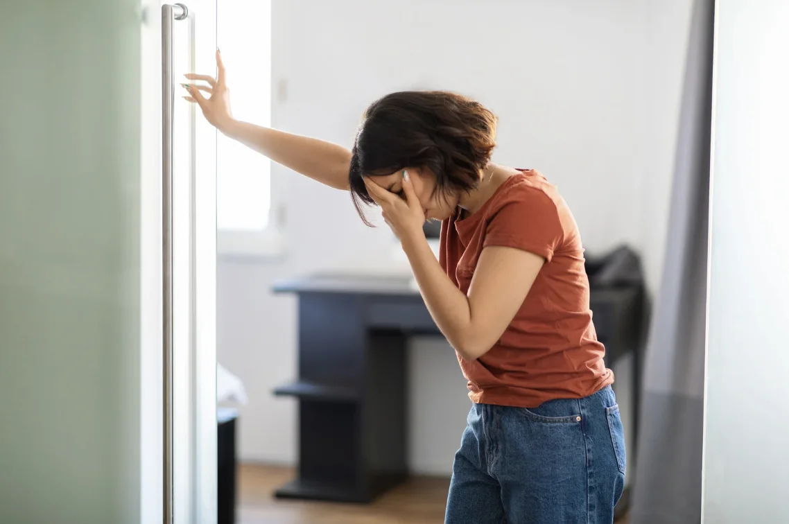 Woman leaning against a doorway holding her head in discomfort, illustrating concerns over whether can dizziness be a sign of eye problems.