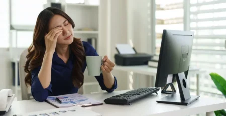 Businesswoman holding a coffee mug and pressing her forehead from digital eye strain in Singapore at her computer desk.
