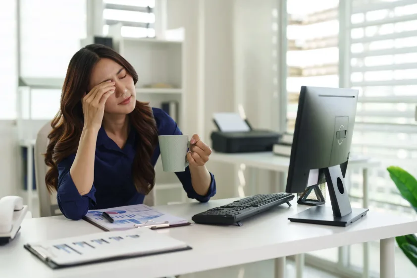 Businesswoman holding a coffee mug and pressing her forehead from digital eye strain in Singapore at her computer desk.